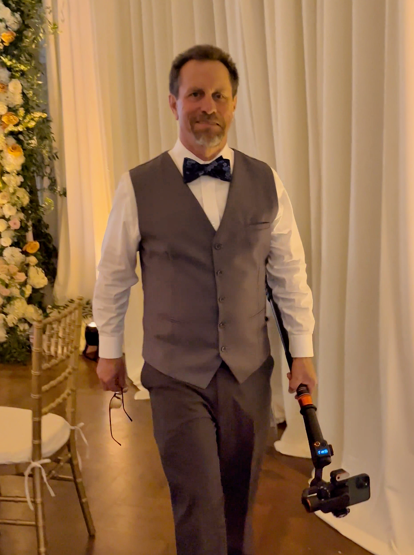 A person wearing a grey vest, white shirt, and bow tie holds a camera stabilizer while walking past white curtains.