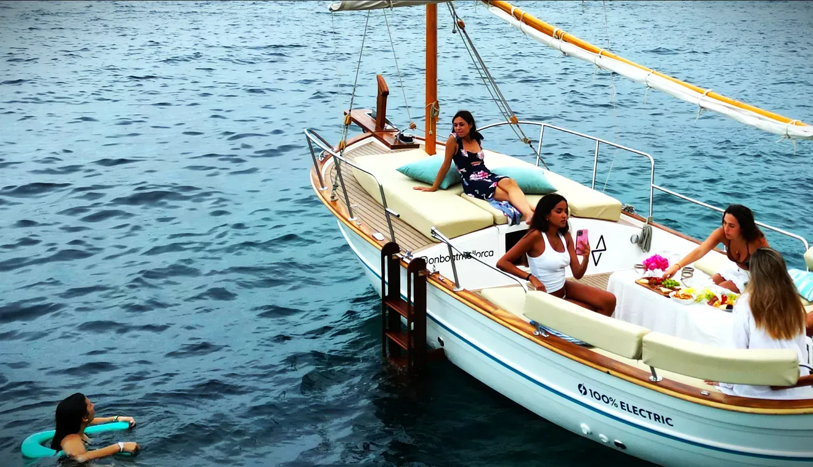 Mujeres descansando en un velero, algunas nadando en el océano. Embarcación blanca con ribetes marrones; día soleado.