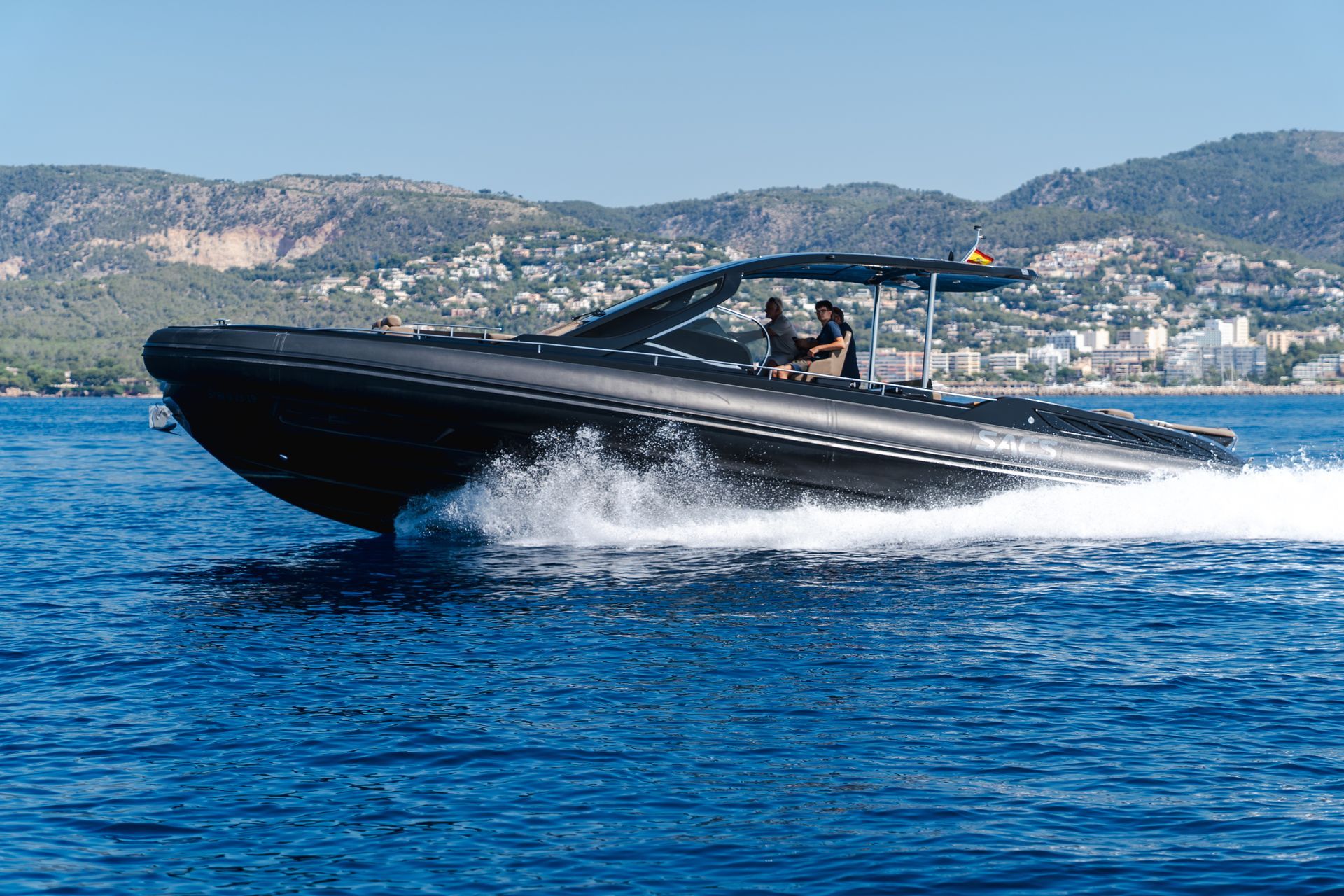 Una lancha negra navega a toda velocidad por aguas azules con montañas al fondo. Dos personas van en la embarcación.