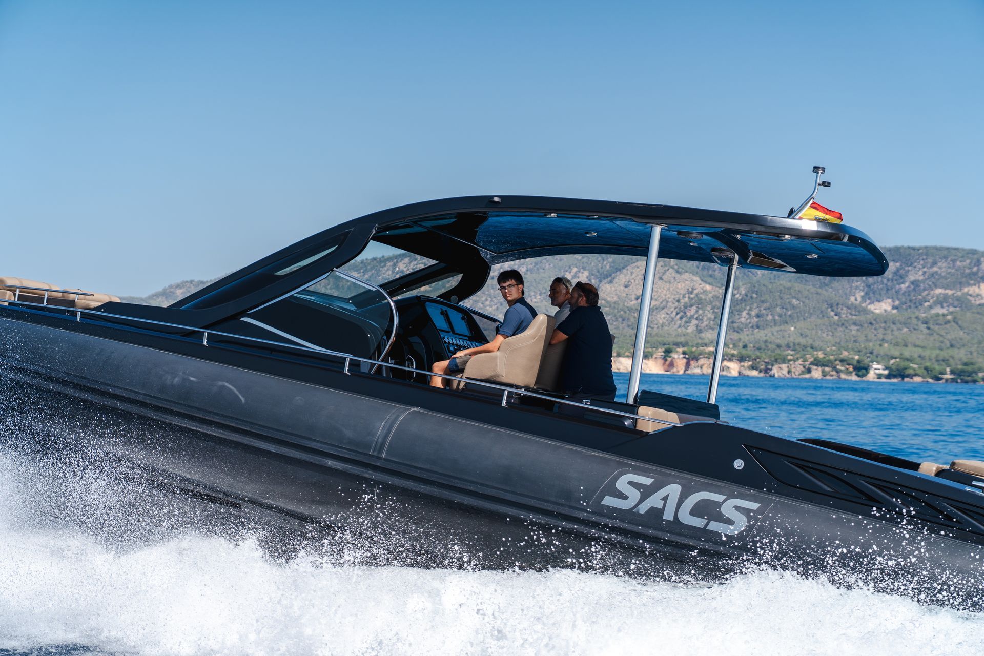 Lancha rápida negra SACS surcando el agua con tres personas a bordo. Cielo azul y costa.