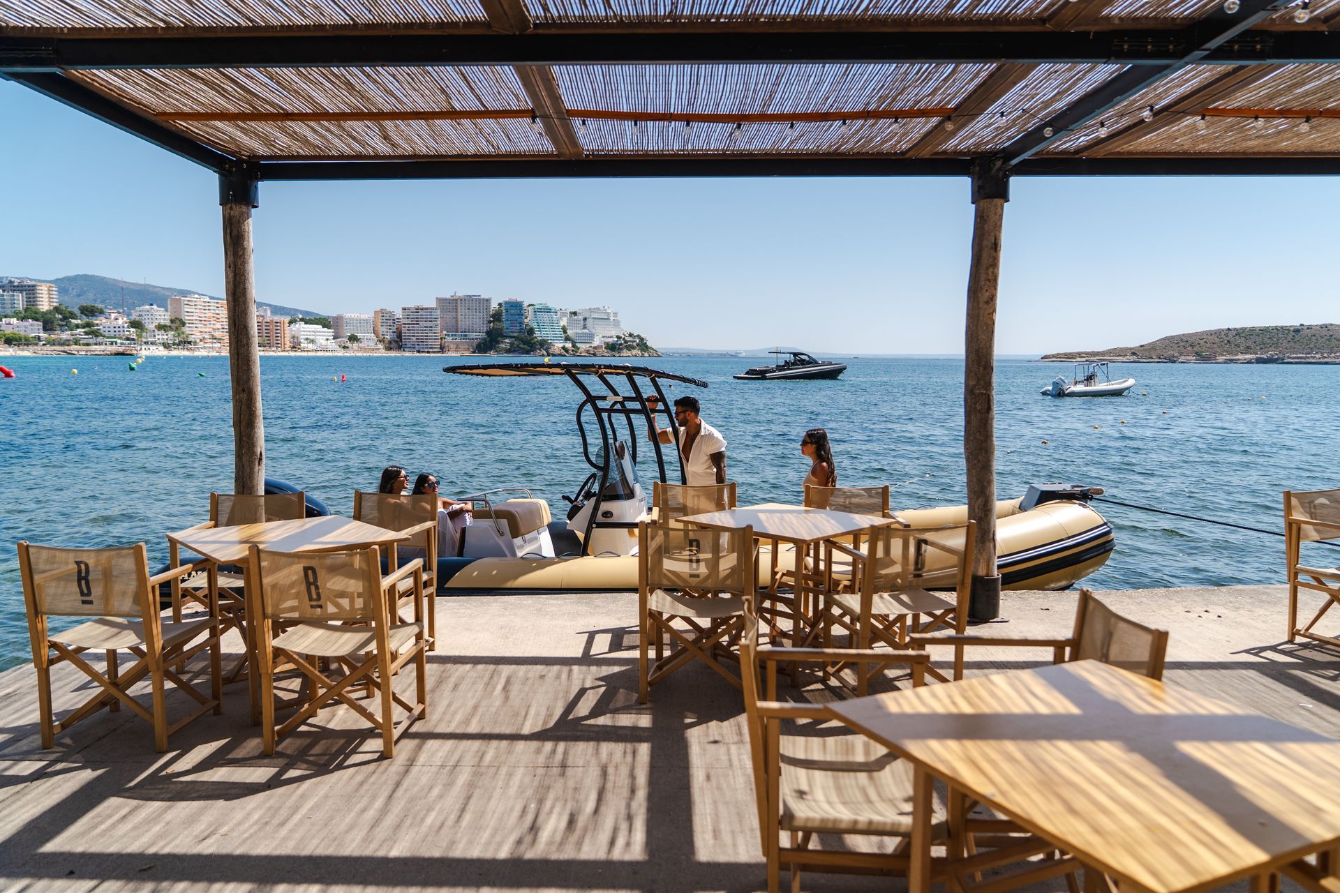 Restaurante al aire libre con mesas y sillas de madera y un muelle con vistas al mar. A lo lejos se ven un barco y edificios.