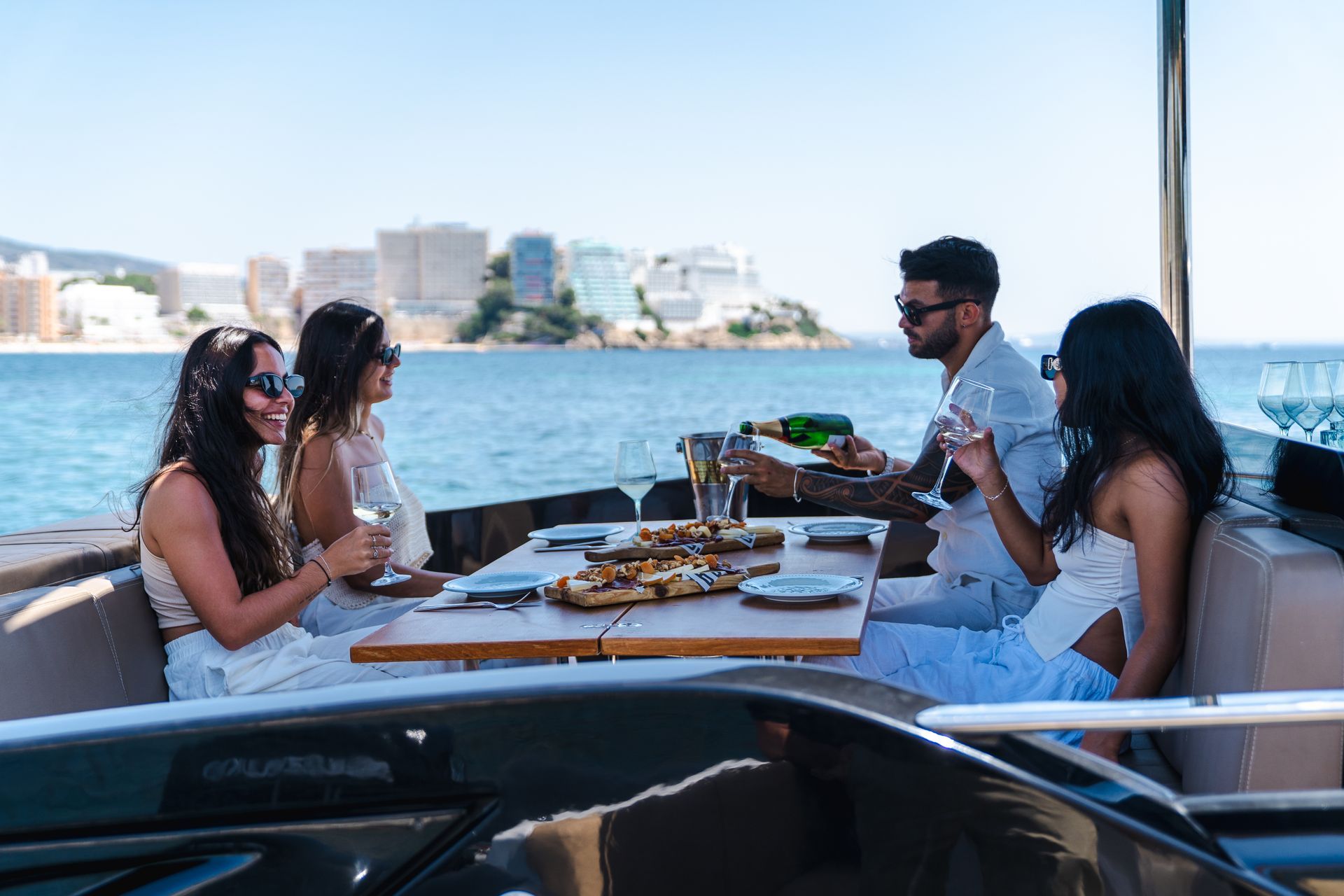 Cuatro personas disfrutan de una comida en un barco junto al mar. Un hombre sirve champán. Agua azul y edificios al fondo.