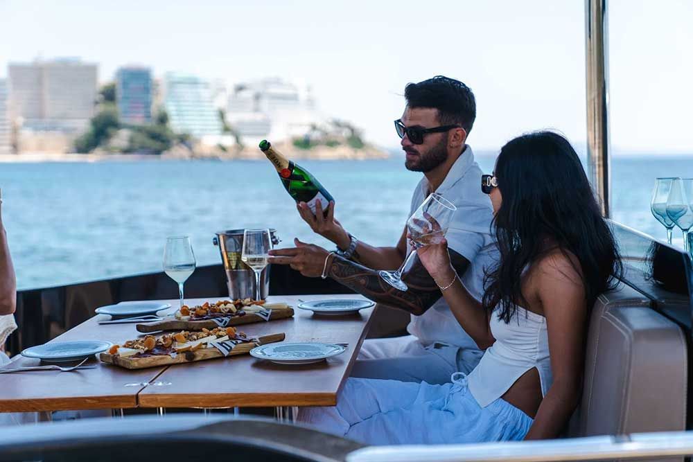 Hombre mostrando una botella de champán a una mujer en un yate. Mesa con comida y vistas al mar.