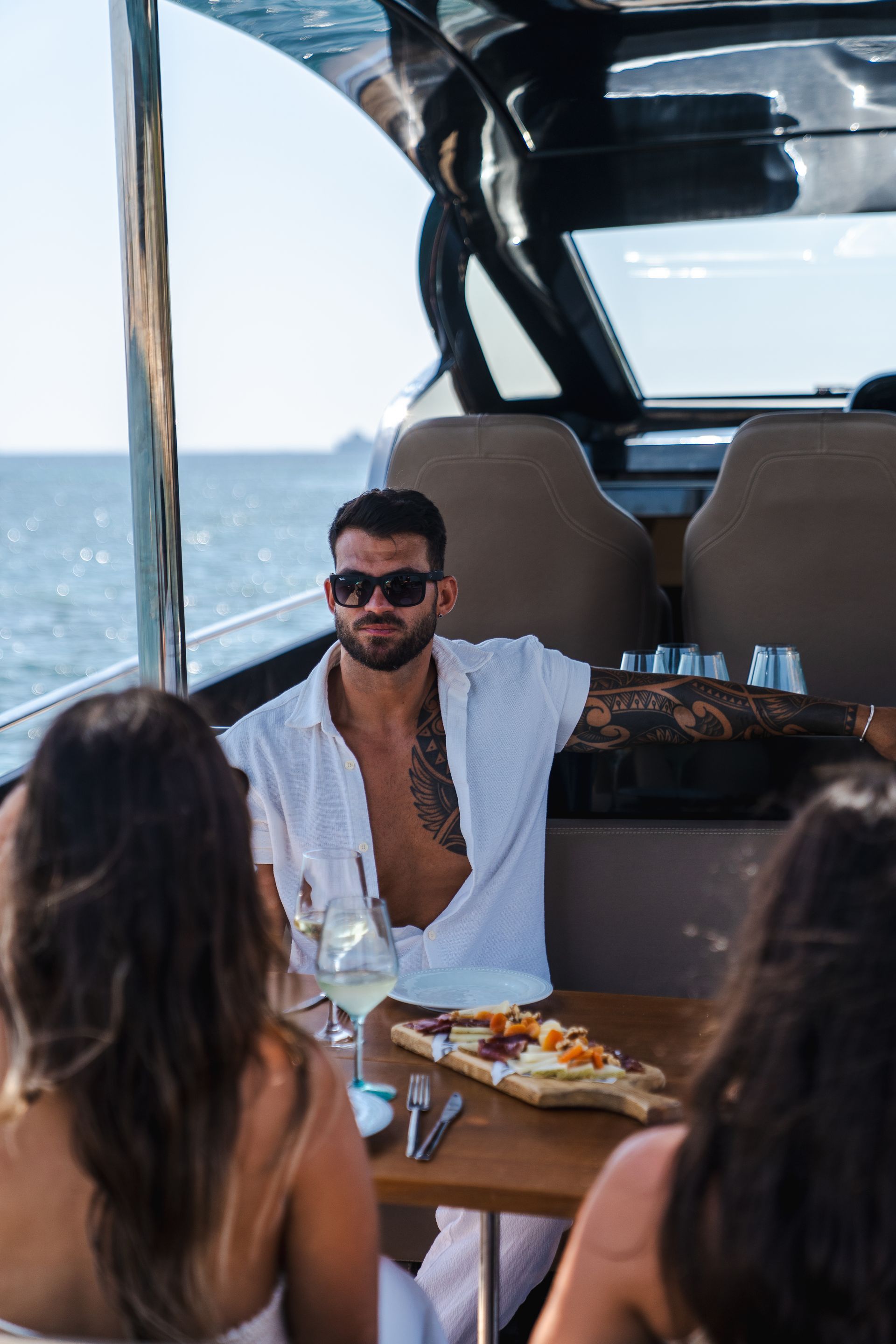 Hombre con gafas de sol en un yate con mujeres, disfrutando de comida y bebida. Día soleado.