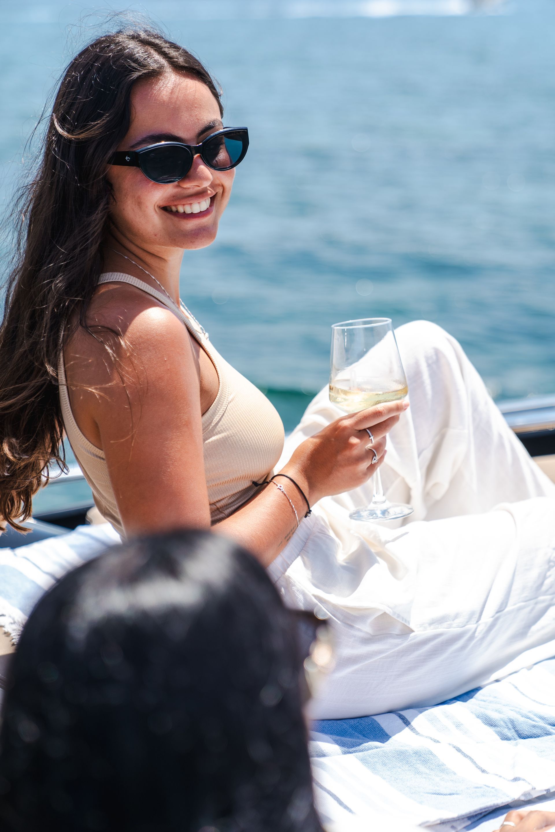 Una mujer con gafas de sol sonríe, sosteniendo una copa de vino, relajándose en un bote cerca del agua.