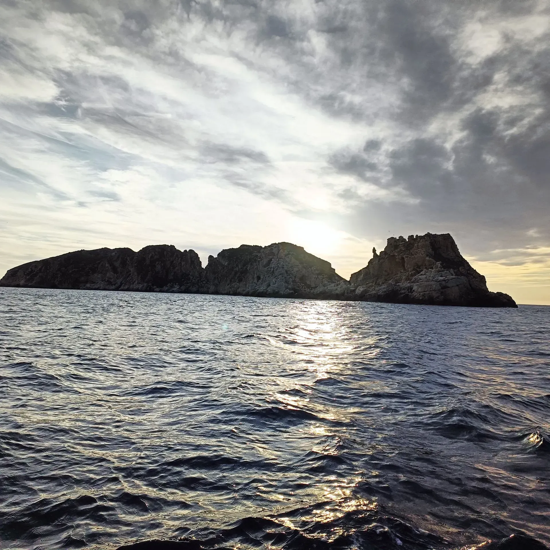 Isla rocosa recortada contra un cielo nublado sobre el océano. El sol se refleja en el agua.