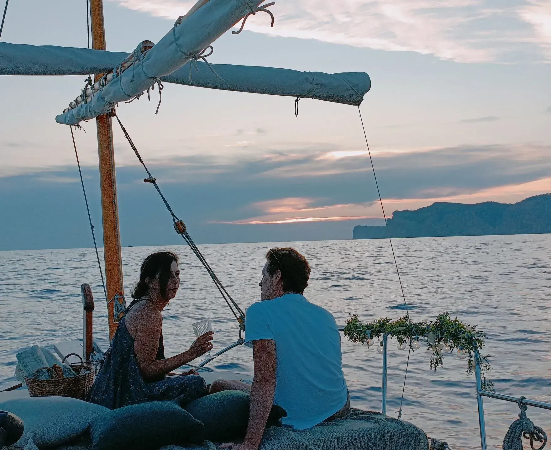 Pareja en un velero al atardecer, sosteniendo bebidas, mirándose, con una montaña al fondo.