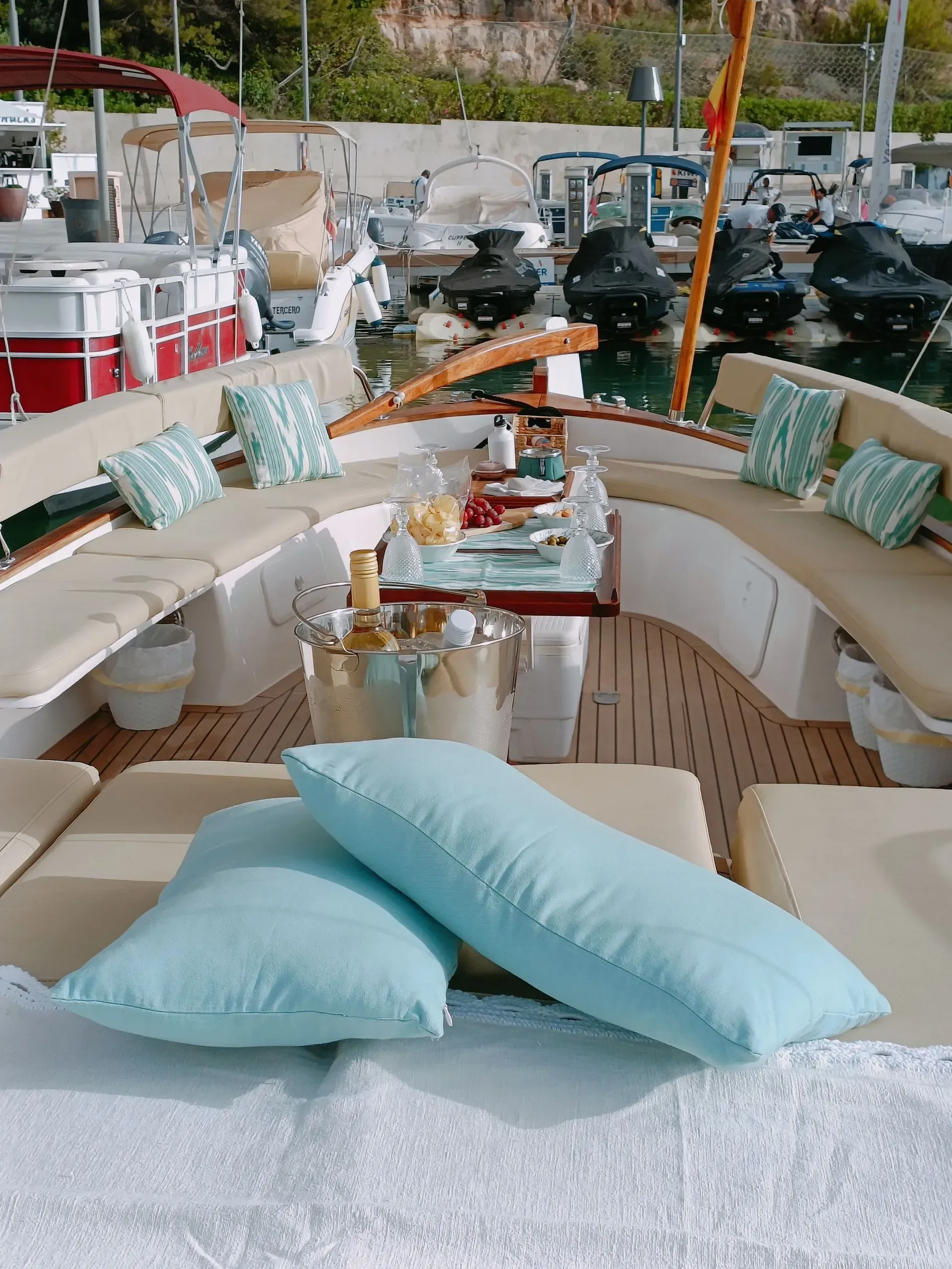 Cubierta de barco con comedor: almohadas de color azul claro, mesa con comida y el puerto al fondo.
