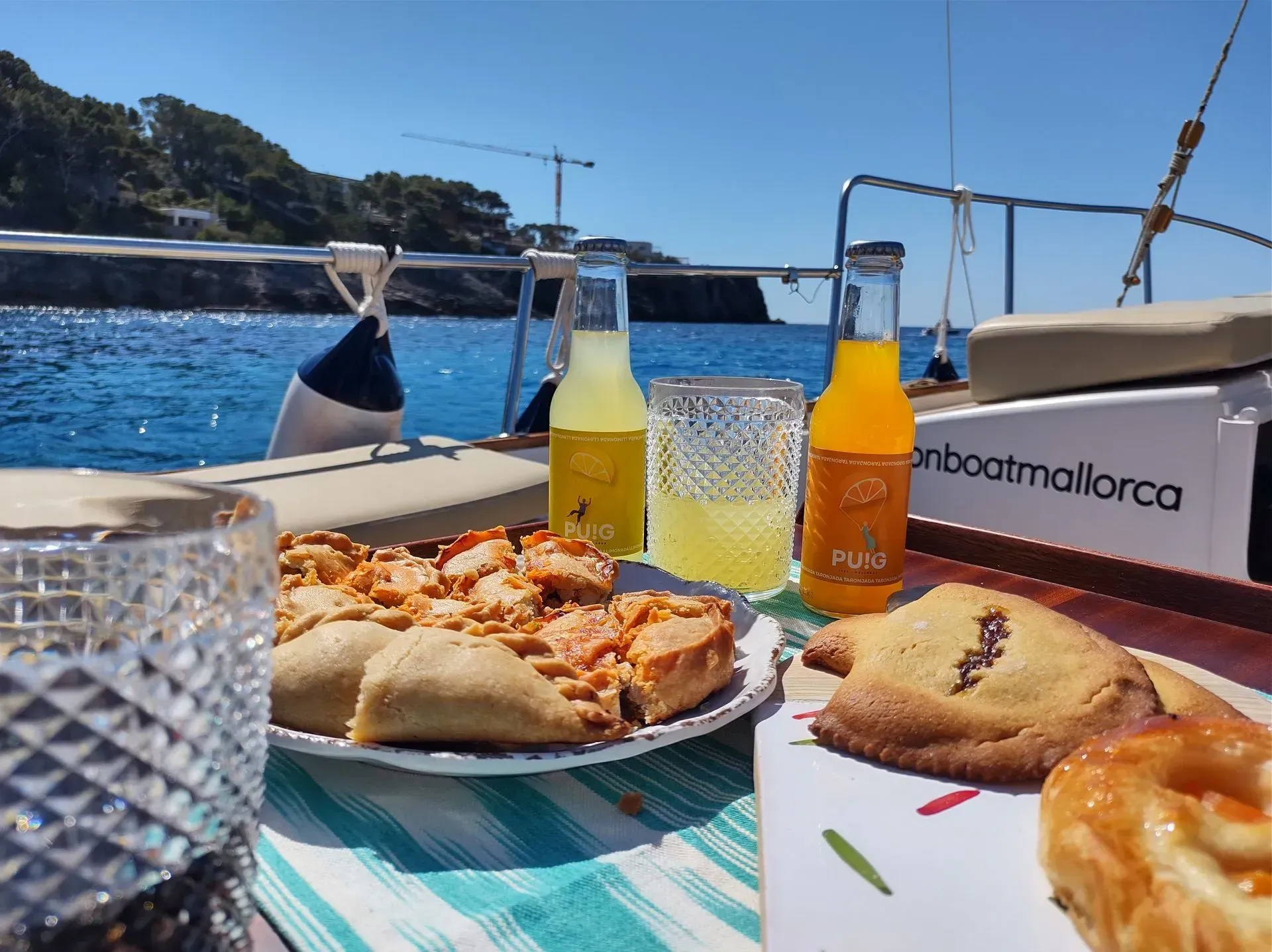 Comida y bebida en un velero: pastel, galletas, botellas de jugo y un vaso, con el mar y el cielo azul de fondo.