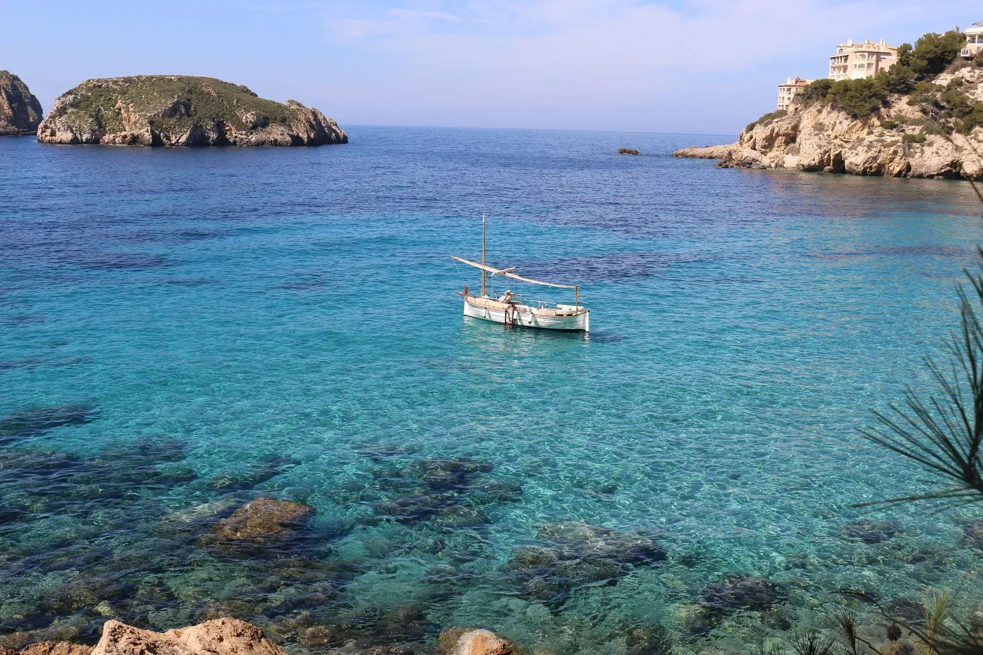 Agua turquesa clara con una pequeña embarcación, una pequeña isla, acantilados rocosos y un día soleado.