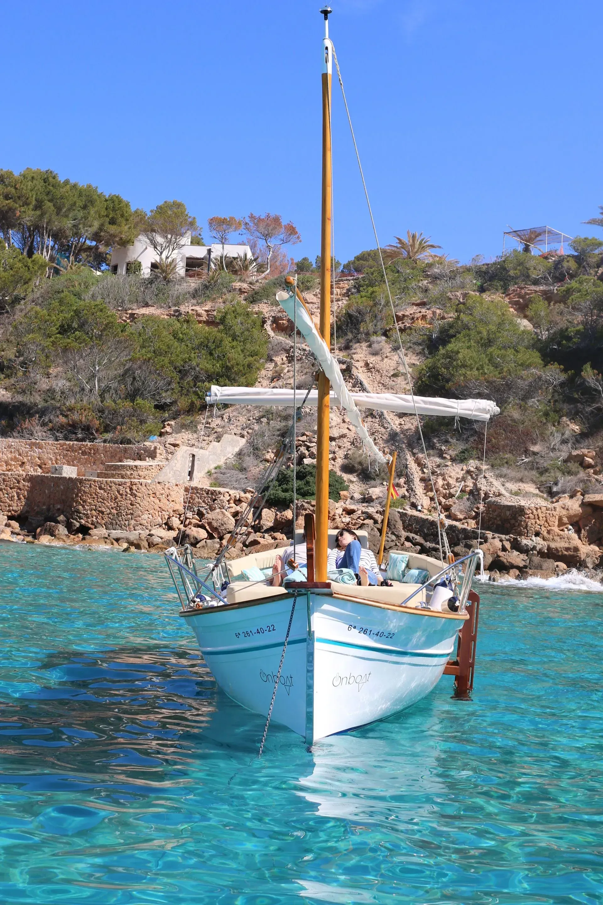 Velero en agua turquesa, mástil de madera, árboles y edificios en la orilla.