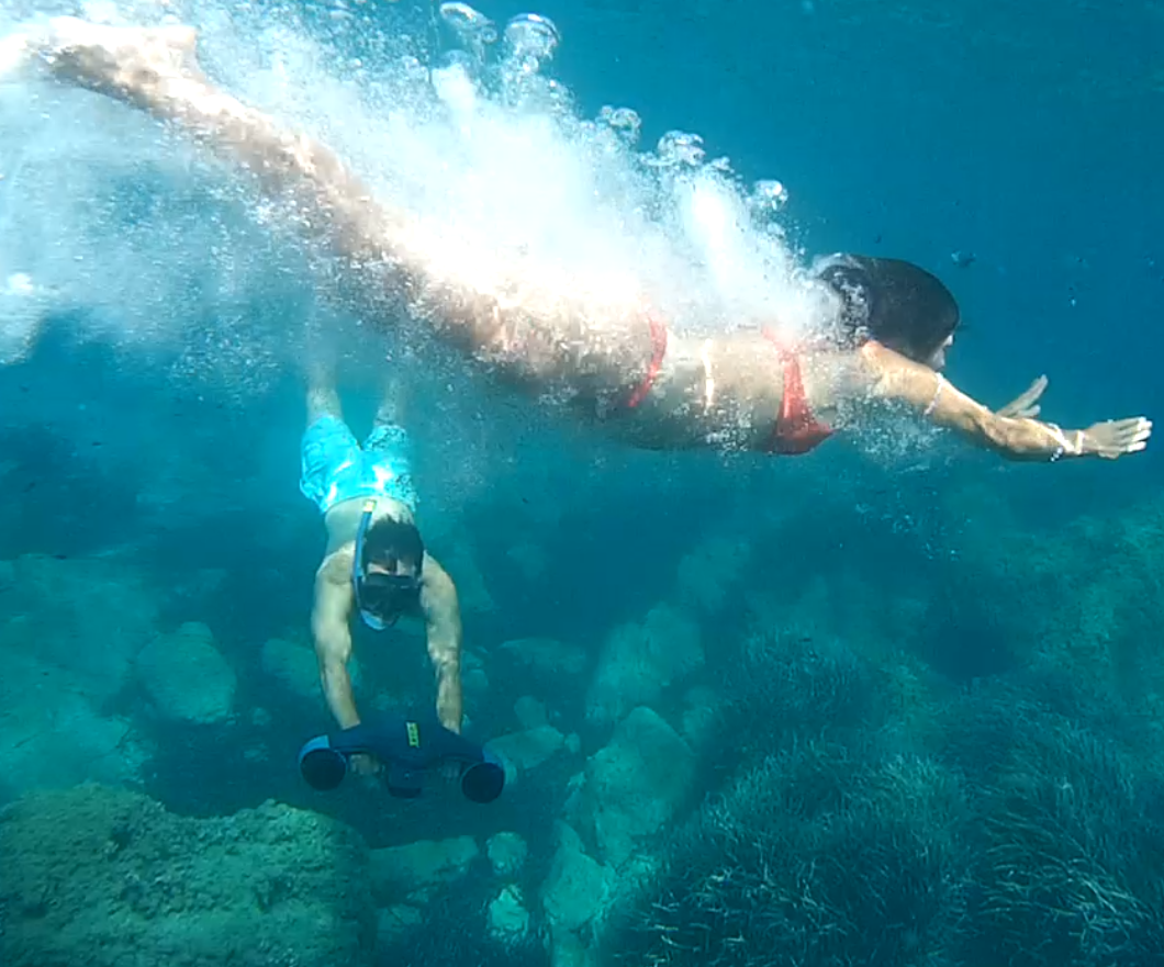Mujer buceando bajo el agua con salpicaduras; hombre haciendo snorkel con un scooter submarino.