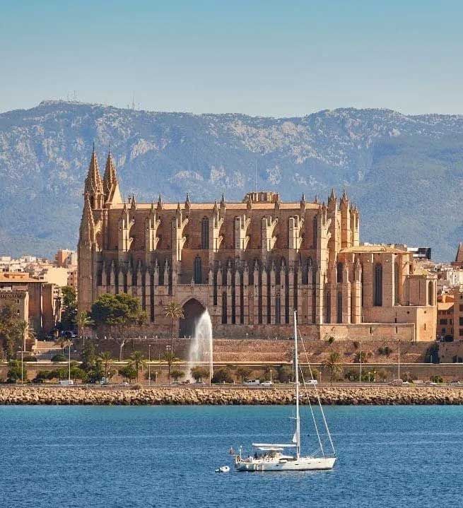 Catedral de Palma de Mallorca, España, con un velero en primer plano, mar y cielo azules, montañas al fondo.