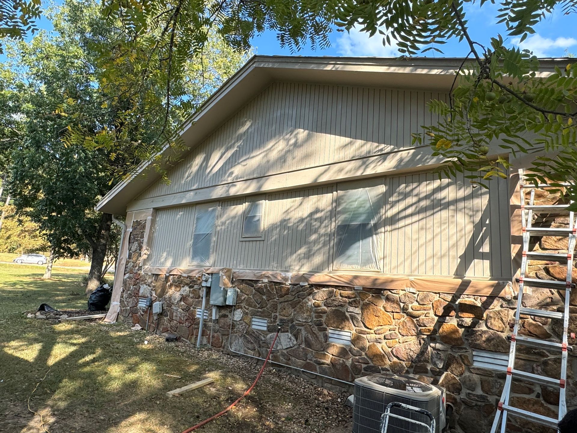 House exterior with stone base, tan siding, and a ladder against the wall. Windows covered. Trees and grass in the background.