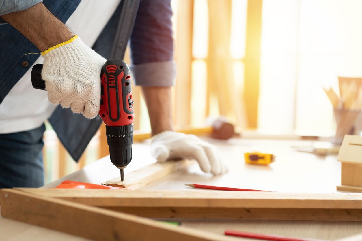 Person wearing gloves using a drill to screw into a wooden frame in a workshop.