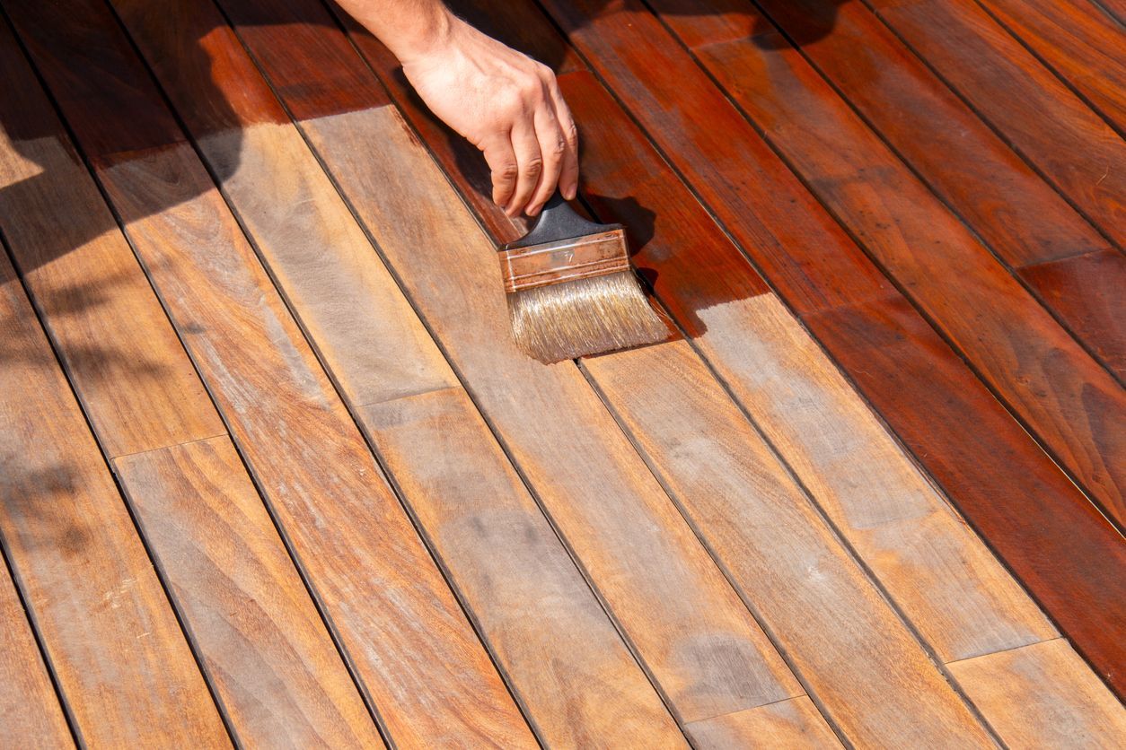 Person staining a wooden deck with a brush. The stained area is dark brown, and the unfinished area is light brown.