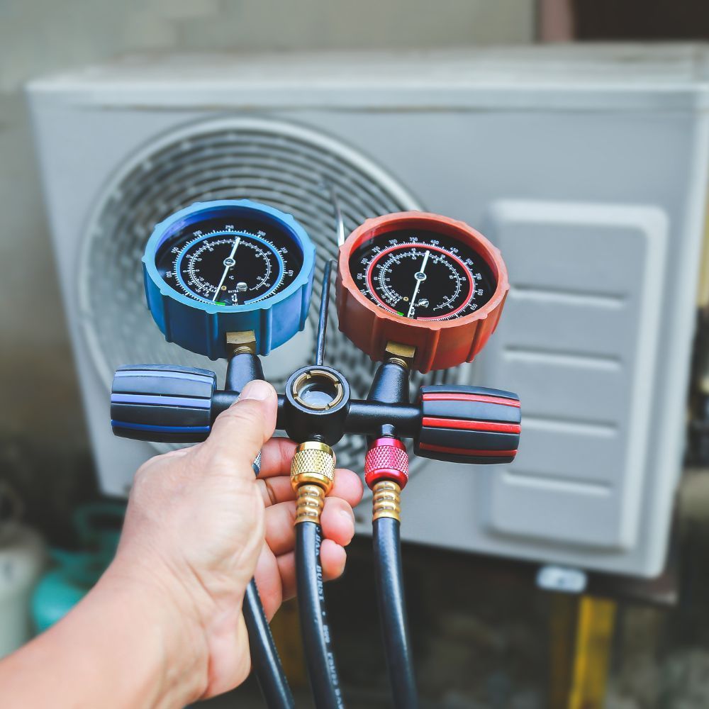 A person is holding two gauges in front of an air conditioner.