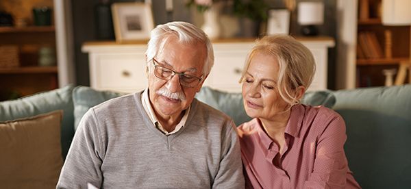 An older couple sitting on a couch, looking at a document. The man is wearing glasses and a sweater, and the woman is in a pink shirt.