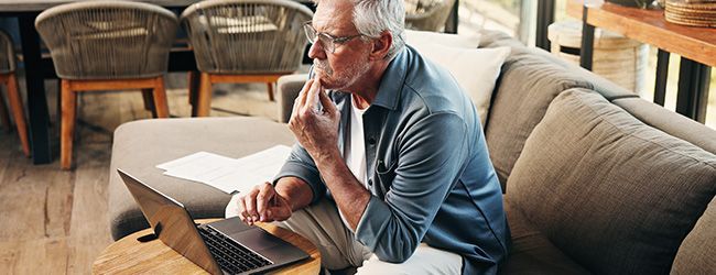Older man sitting on a couch, looking at a laptop and holding a phone in a living room