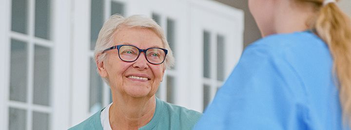 An elderly woman wearing glasses is smiling while talking to a nurse.