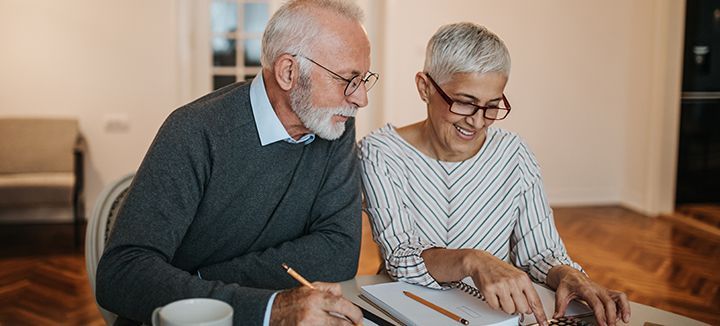 An elderly couple is sitting at a table looking at a laptop.