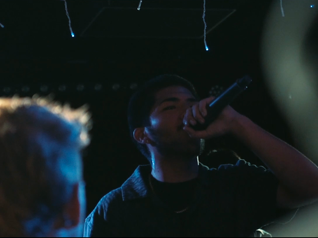 Man singing into a microphone on stage, under blue lights.