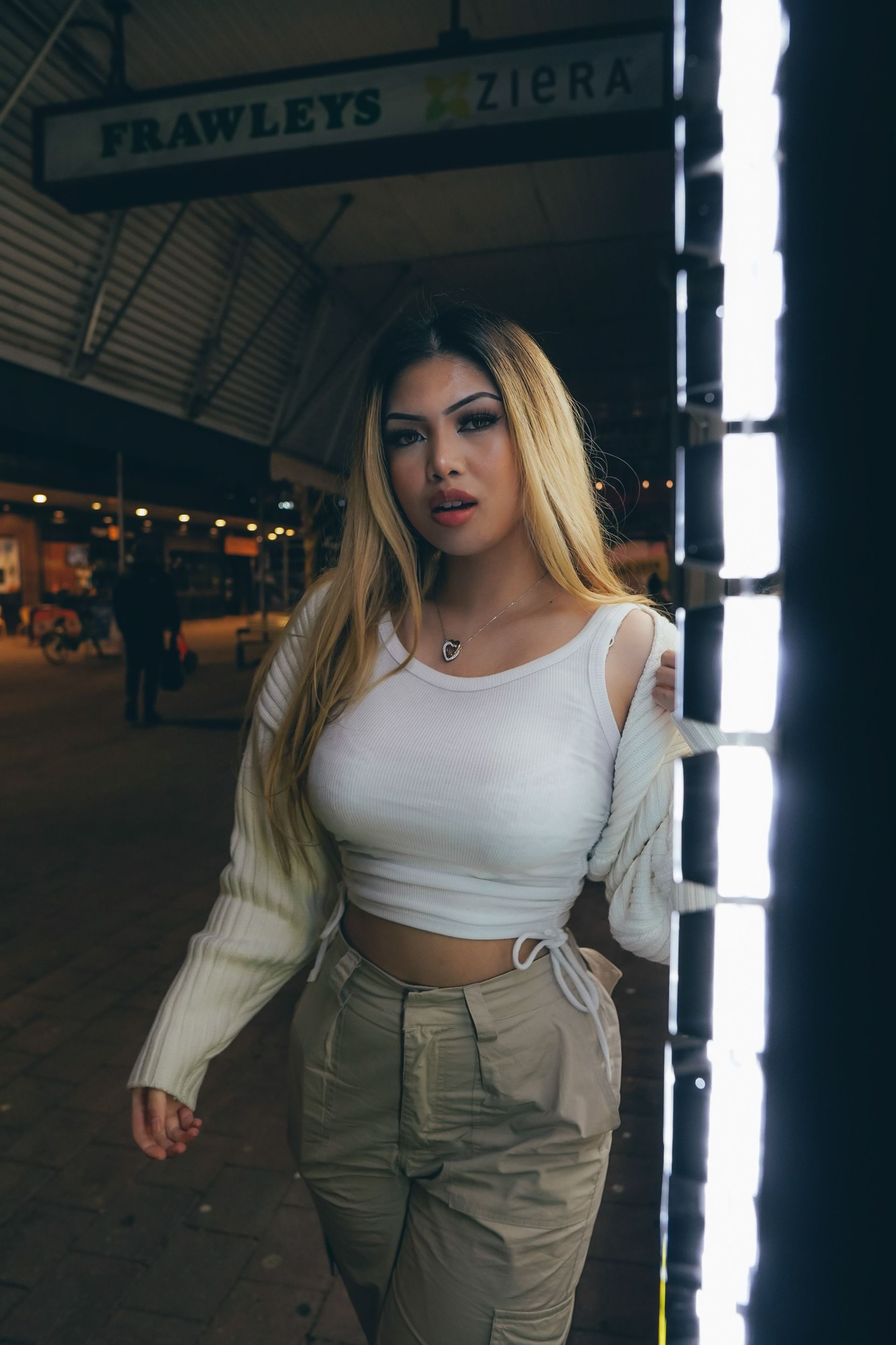 Woman in white top and tan pants, poses by a bright light at night.