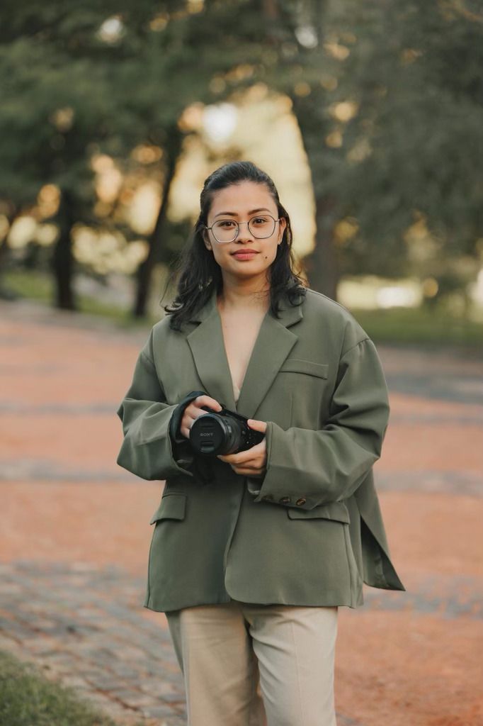 Woman leans out car window, brown top, gazing. White car, city background, arms on ledge.