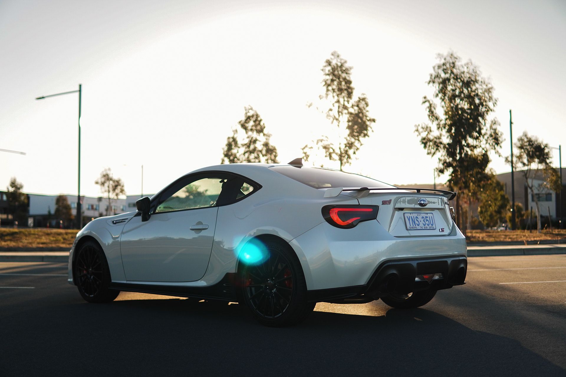 White sports car parked on asphalt, near trees and buildings, with the sun shining.