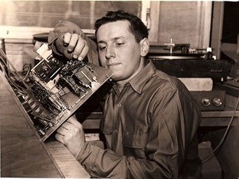 Man examines electronic circuit board, indoors. Wearing a button-down shirt, turntable in background.