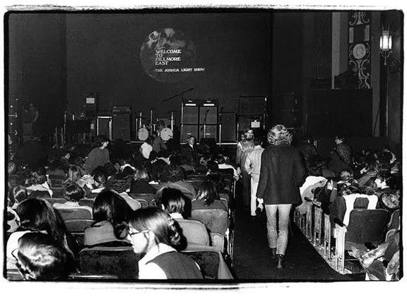 Audience at a concert in a theater. Stage features band equipment. A person walks in aisle.