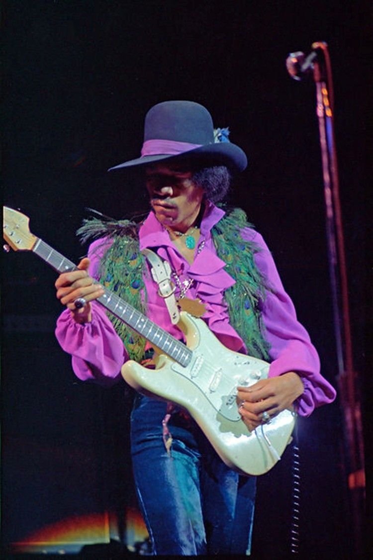 Jimi Hendrix playing a white guitar on stage, wearing a purple shirt, hat, and a green feathered vest.