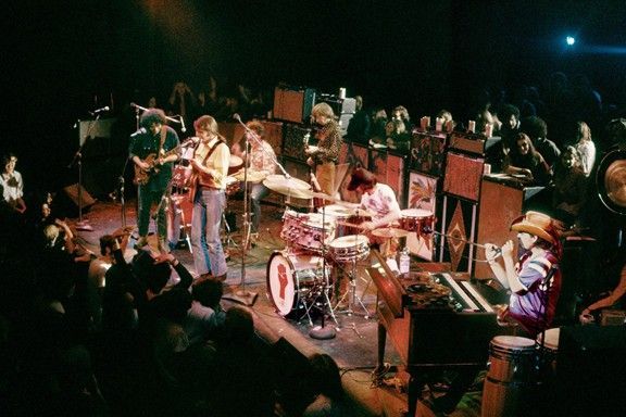 Band on stage, playing instruments; audience watching. Dark setting, bright lights.
