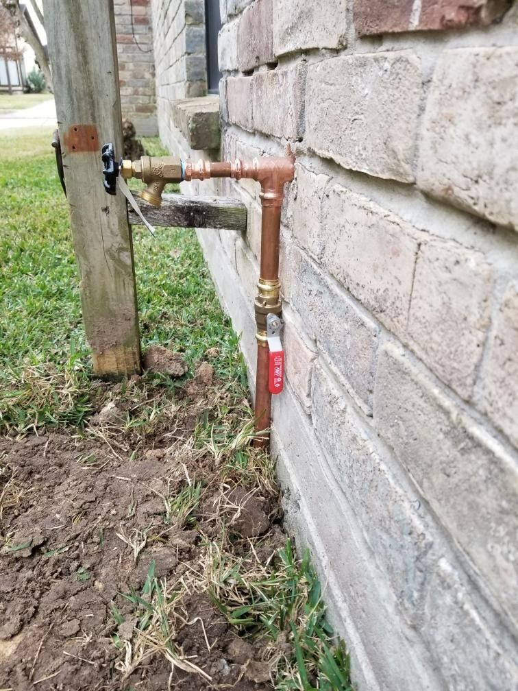 Copper pipes and valves attached to a brick wall. A spigot is near a wooden post.