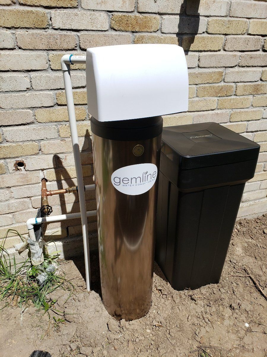 Water softener system installed outdoors against a brick wall. A brown tank is next to a black salt container.