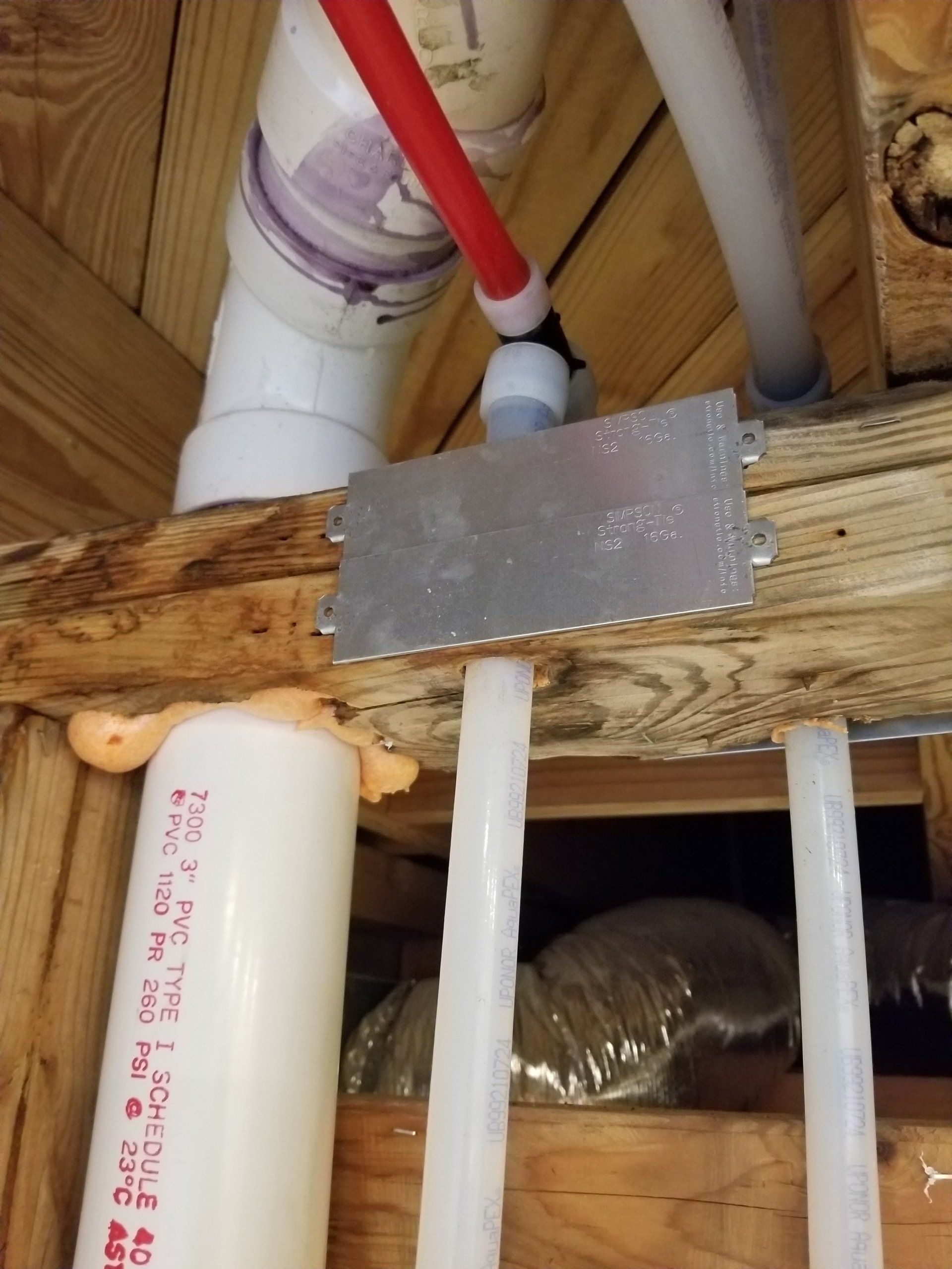 Pipes, including white PVC and red PEX, are attached to a wooden beam; a metal plate secures them.