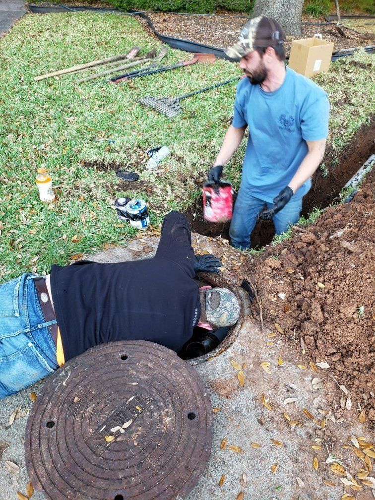 Two men working on a manhole in a yard. One leans in, another pours from a can. Dirt and tools surround them.
