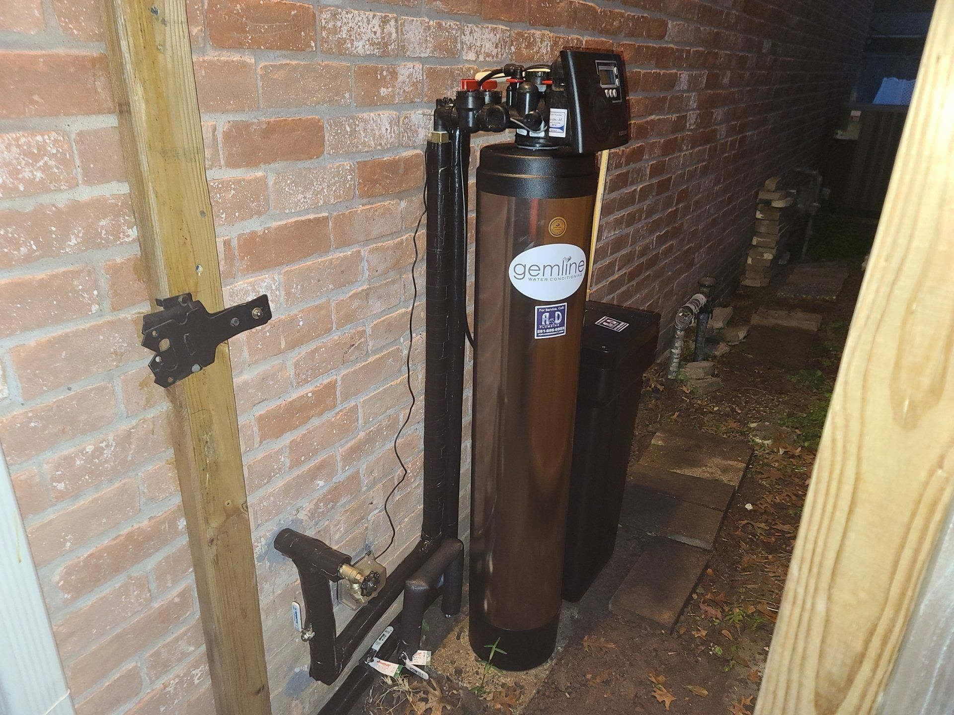 Water softener system installed against a brick wall. Black pipes connect to a brown tank with digital controls.