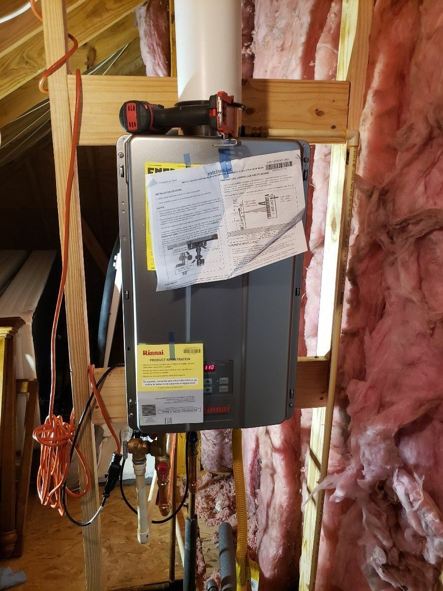 Gray and black tankless water heater mounted in an attic, with insulation and wood framing visible.