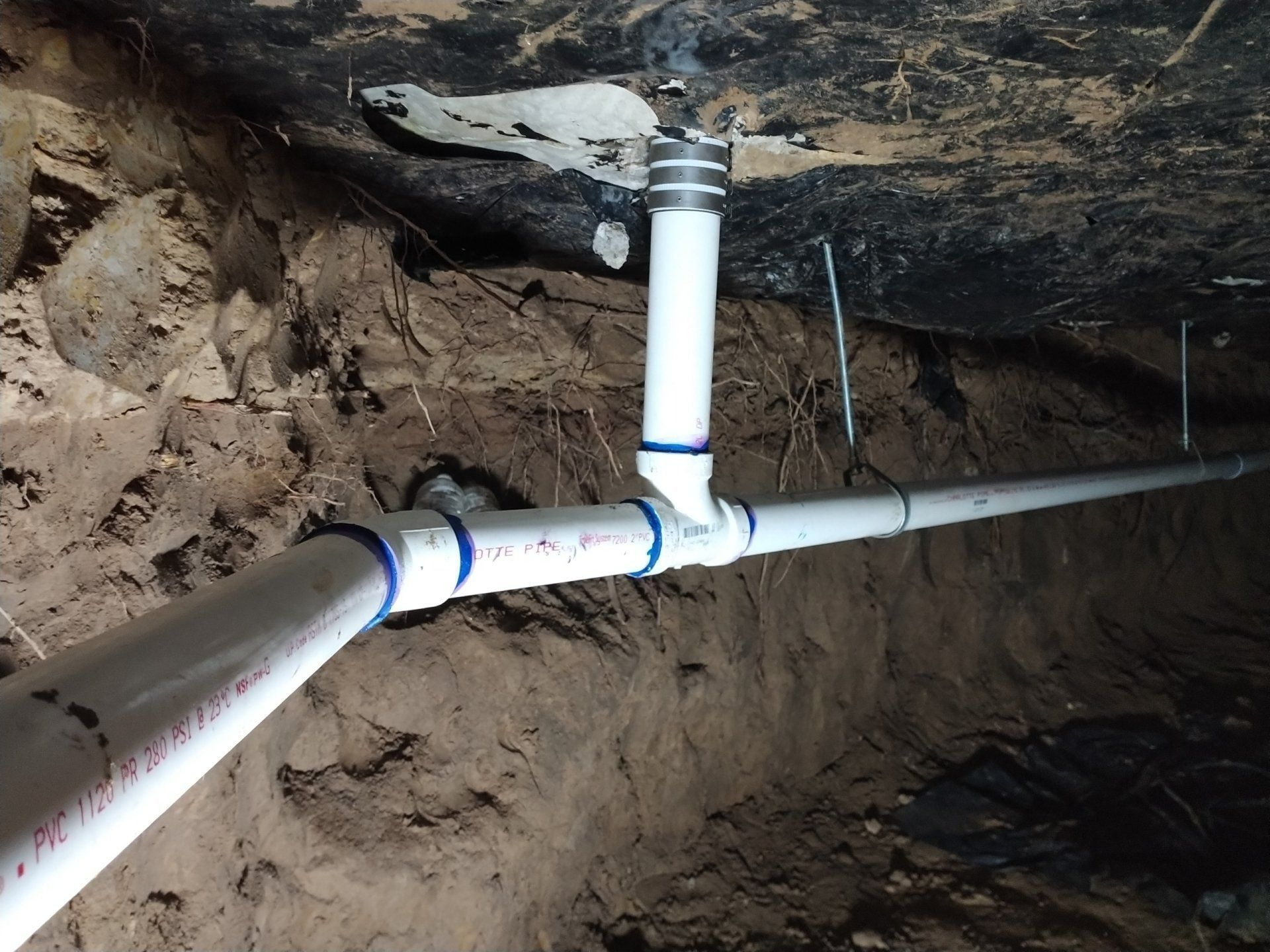 White PVC pipes with sprinkler head and connections in a dark earthen trench.
