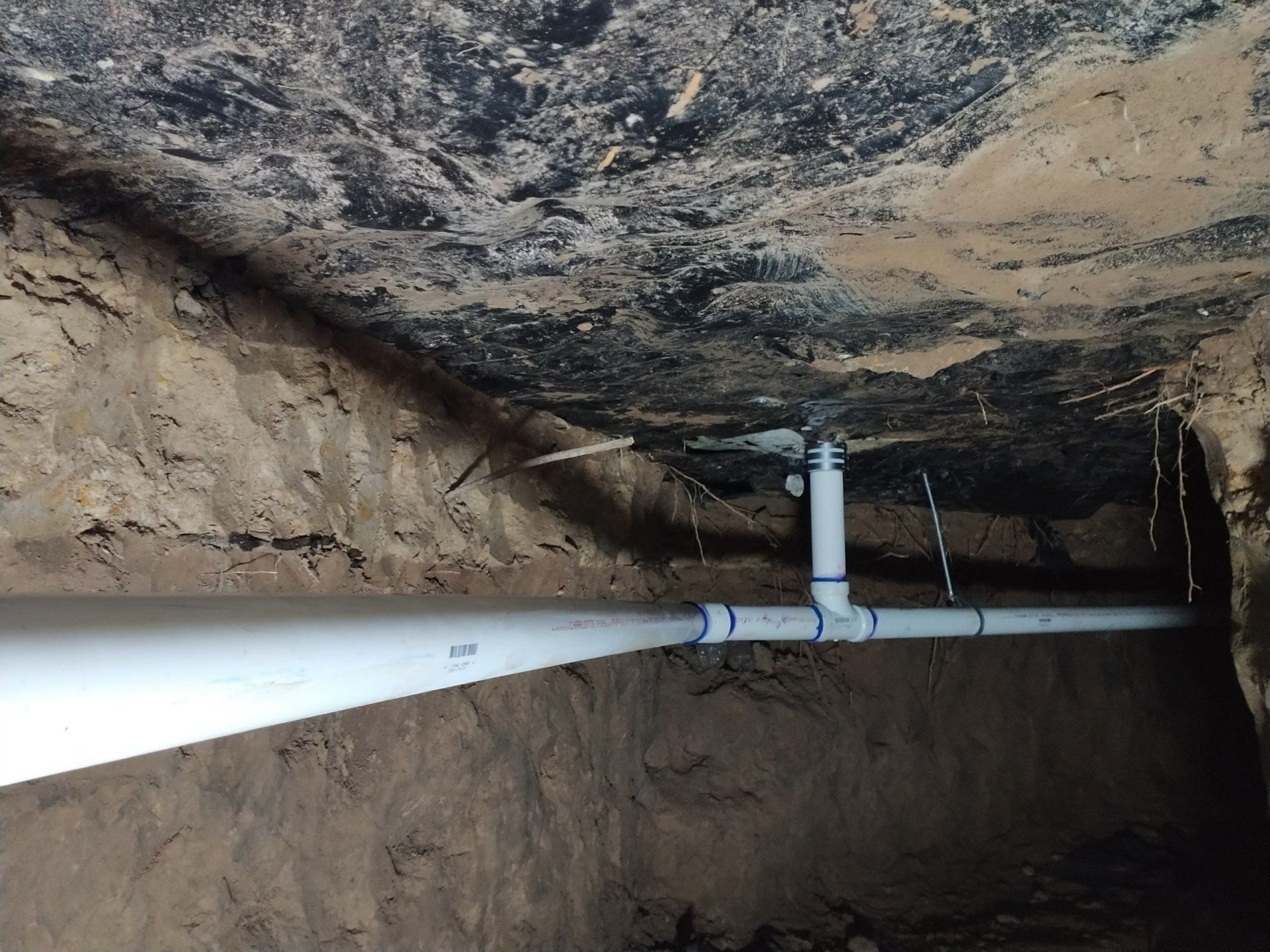 White PVC pipes in a trench under a dark ceiling. Joints and fittings visible, soil walls.