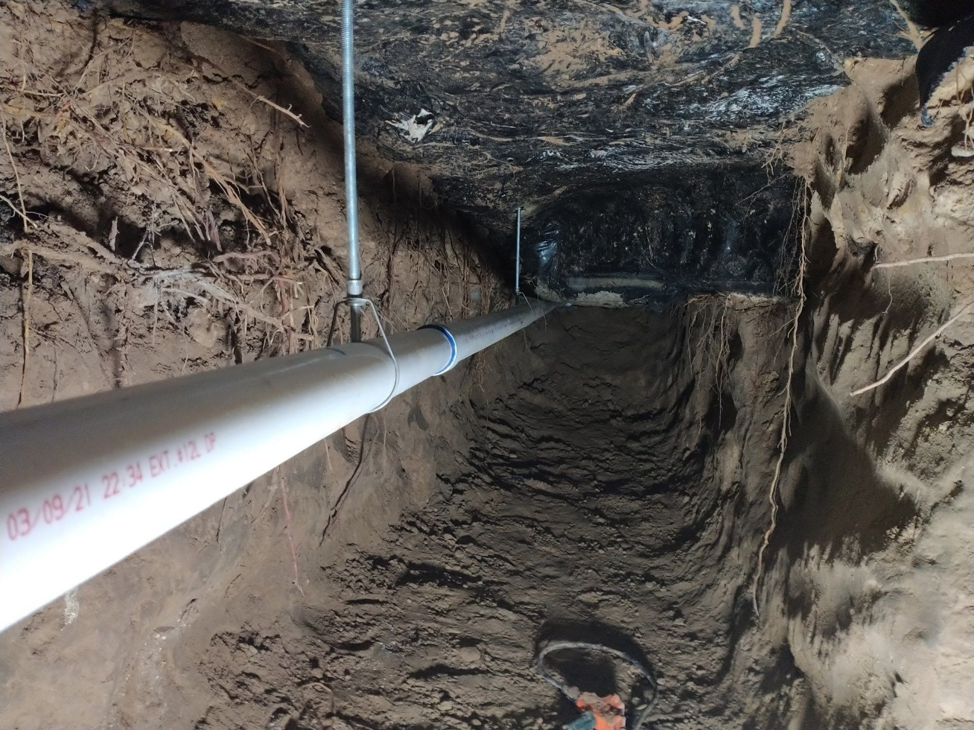 White PVC pipe in a narrow trench. The trench is dug in earth beneath a dark, low ceiling.
