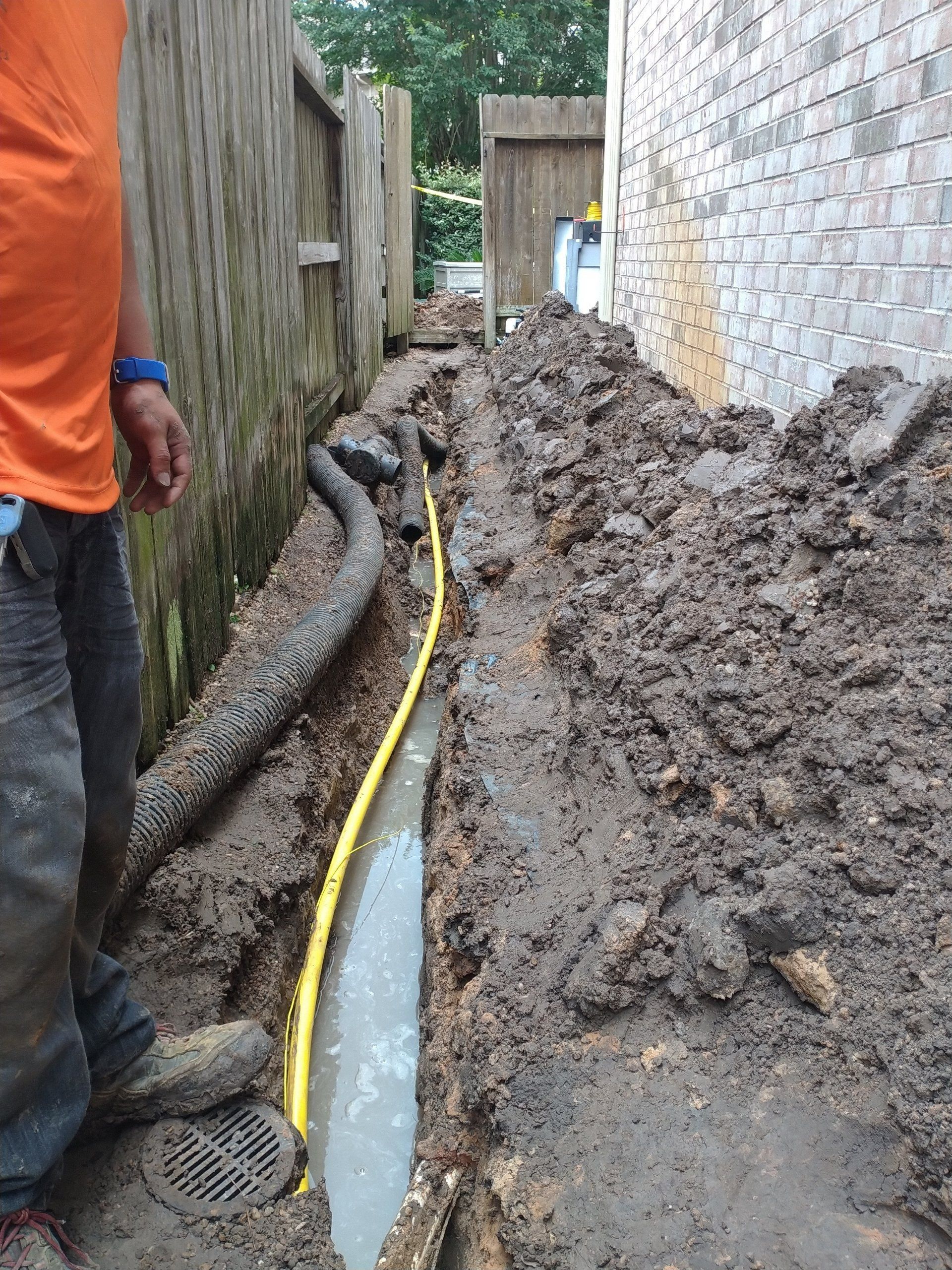 Ditch with water, yellow pipe, and a black corrugated pipe; next to a fence and a brick wall; person in orange shirt.