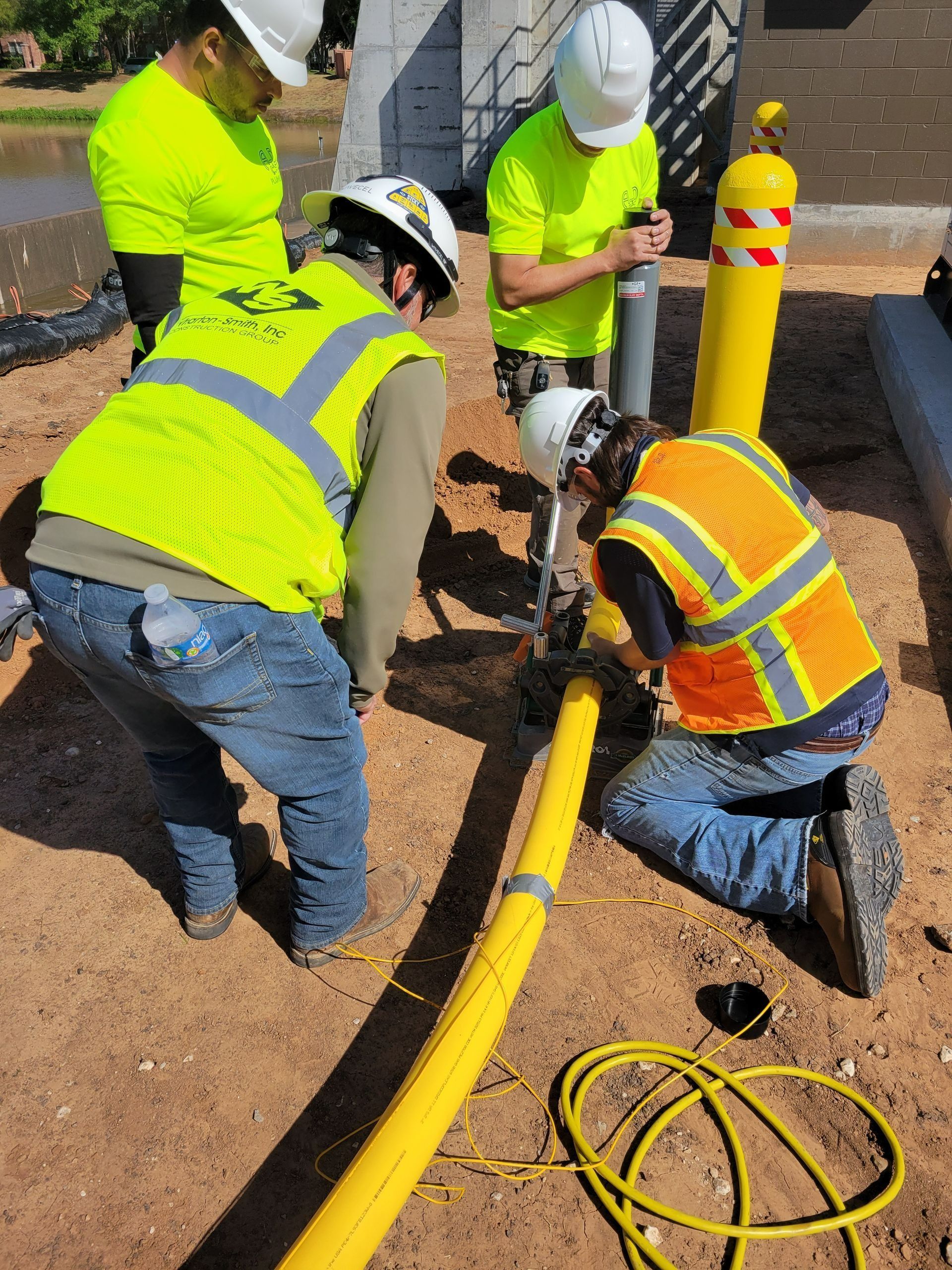 Construction workers in neon vests install a yellow pipe outdoors.