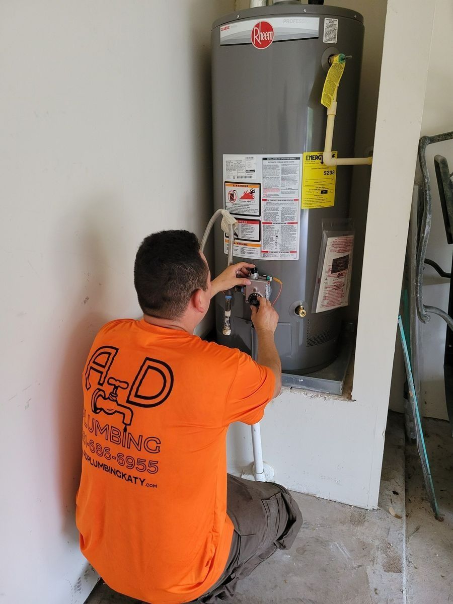 Plumber in orange shirt working on a tall, gray water heater in a corner.