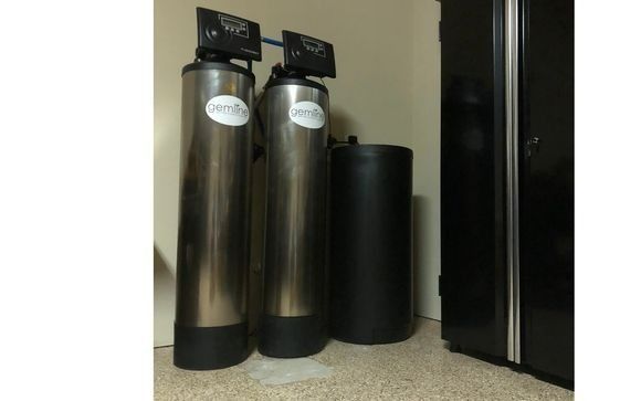 Two tall, silver water treatment tanks with black components next to a black cabinet on carpet.