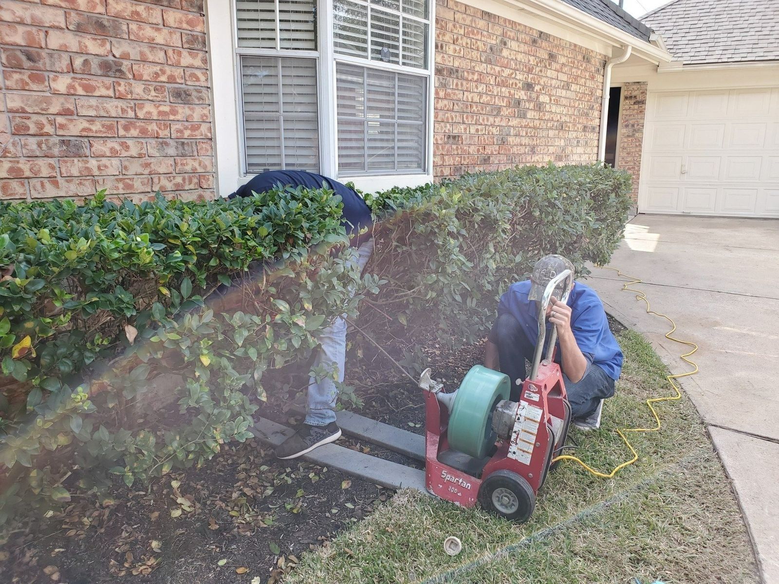 Two people clearing a drain near a brick house, using a red drain cleaning machine.