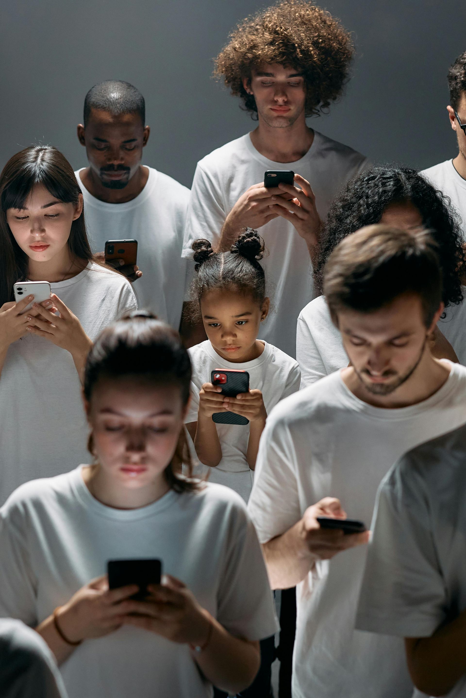People in white t-shirts are fixated on their smartphones, lit by a dim light.
