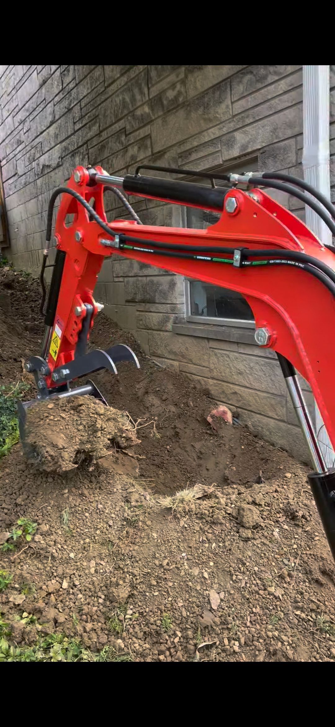 A red mini excavator digs a trench next to a weathered stone wall.