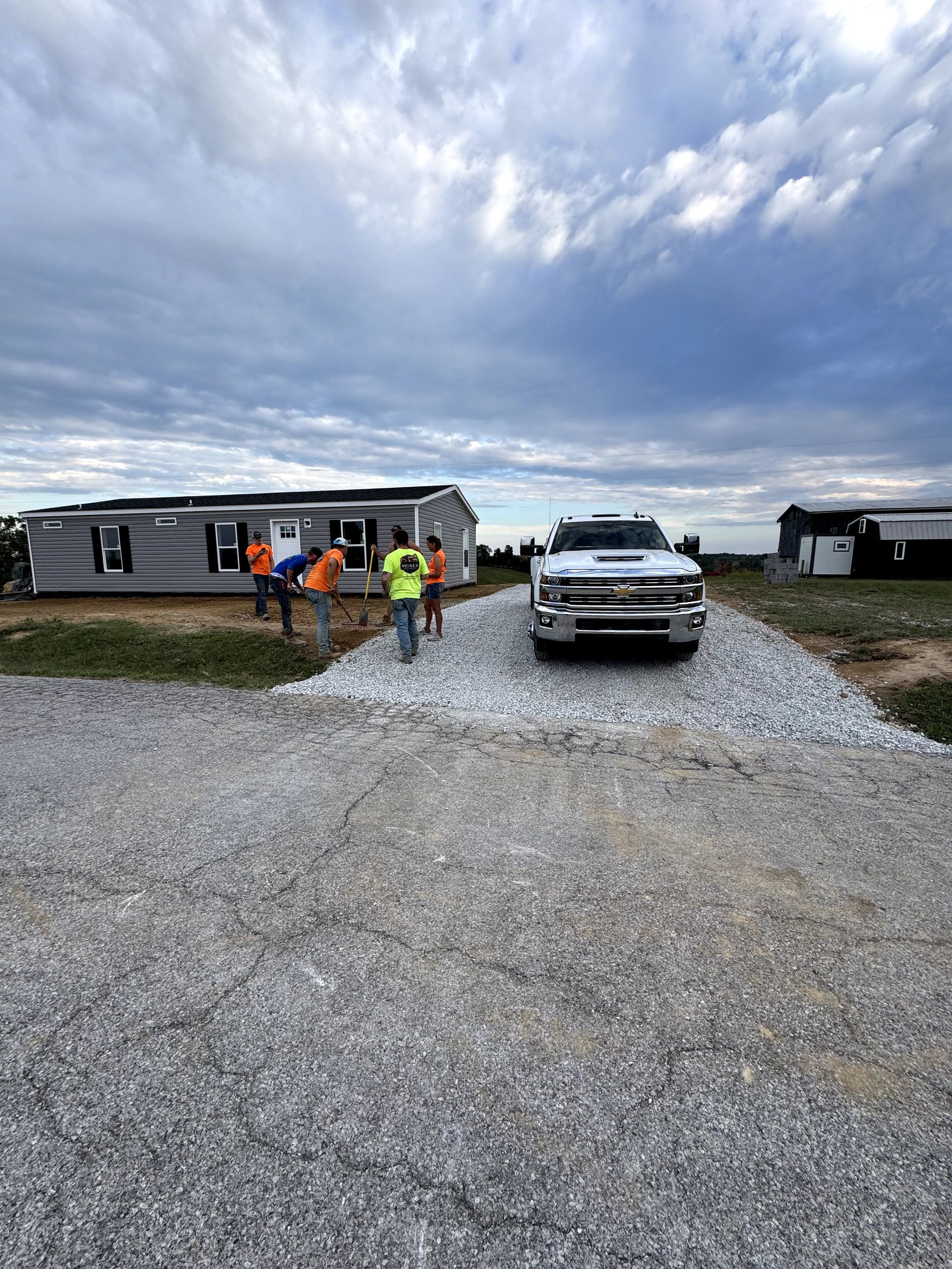 Workers installing gravel driveway near a gray mobile home and white truck under cloudy sky.